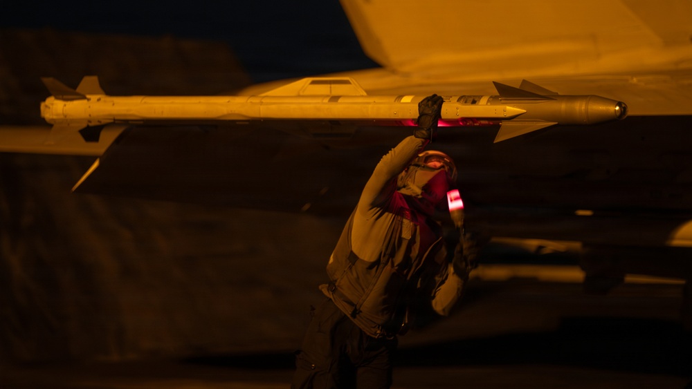 A military technician or ordnance crew member is working on an aircraft at night, handling what appears to be an air-to-air missile mounted under the wing. The scene is illuminated by warm amber/orange artificial lighting, creating a dramatic low-light environment. The worker appears to be inspecting or securing the missile to the aircraft's wing pylon.