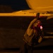 A military technician or ordnance crew member is working on an aircraft at night, handling what appears to be an air-to-air missile mounted under the wing. The scene is illuminated by warm amber/orange artificial lighting, creating a dramatic low-light environment. The worker appears to be inspecting or securing the missile to the aircraft's wing pylon.