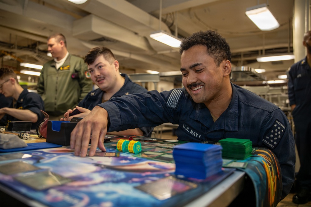 U.S. Navy personnel are playing a trading card game, likely Magic: The Gathering, in what appears to be a common area aboard a naval vessel. The sailors are in uniform and appear relaxed and engaged in recreational activity during off-duty time. The setting features industrial overhead piping and fluorescent lighting typical of a ship's interior.