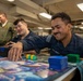 U.S. Navy personnel are playing a trading card game, likely Magic: The Gathering, in what appears to be a common area aboard a naval vessel. The sailors are in uniform and appear relaxed and engaged in recreational activity during off-duty time. The setting features industrial overhead piping and fluorescent lighting typical of a ship's interior.