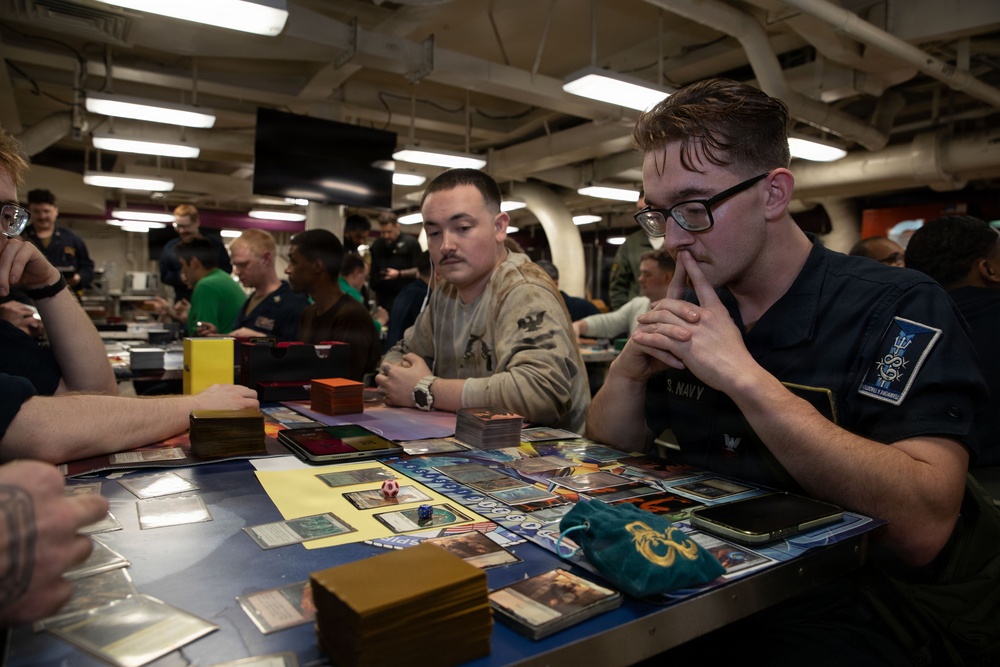 A group of U.S. Navy personnel are gathered in what appears to be a shipboard common area or mess deck, playing a trading card game, most likely Magic: The Gathering. Two primary players are visible in the foreground, with one wearing a U.S. Navy uniform patch, both deeply focused on the game. The background shows numerous other service members engaged in similar tabletop gaming activities.