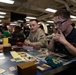 A group of U.S. Navy personnel are gathered in what appears to be a shipboard common area or mess deck, playing a trading card game, most likely Magic: The Gathering. Two primary players are visible in the foreground, with one wearing a U.S. Navy uniform patch, both deeply focused on the game. The background shows numerous other service members engaged in similar tabletop gaming activities.
