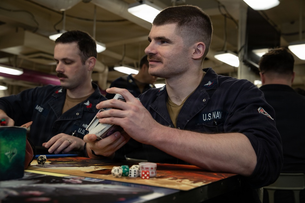Two U.S. Navy sailors in dark blue utility uniforms are seated at a table playing a tabletop card and dice game, likely Magic: The Gathering or a similar trading card game. The foreground sailor is holding a deck of cards while multiple dice of various colors are visible on the game mat. The setting appears to be an interior compartment aboard a naval vessel, indicated by the industrial ceiling with fluorescent lighting and pipes.