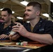 Two U.S. Navy sailors in dark blue utility uniforms are seated at a table playing a tabletop card and dice game, likely Magic: The Gathering or a similar trading card game. The foreground sailor is holding a deck of cards while multiple dice of various colors are visible on the game mat. The setting appears to be an interior compartment aboard a naval vessel, indicated by the industrial ceiling with fluorescent lighting and pipes.