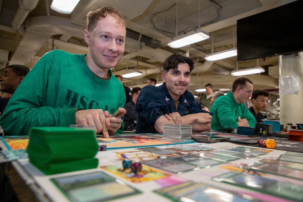Two young men are seated at a table playing a trading card game, likely Magic: The Gathering, with cards spread across the playing surface. The setting appears to be an indoor facility with industrial-style ceiling lighting, possibly a military vessel or base given the visible 'HSC' text on one player's sweatshirt. Additional players are visible in the background engaged in similar tabletop gaming activities.