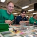 Two young men are seated at a table playing a trading card game, likely Magic: The Gathering, with cards spread across the playing surface. The setting appears to be an indoor facility with industrial-style ceiling lighting, possibly a military vessel or base given the visible 'HSC' text on one player's sweatshirt. Additional players are visible in the background engaged in similar tabletop gaming activities.