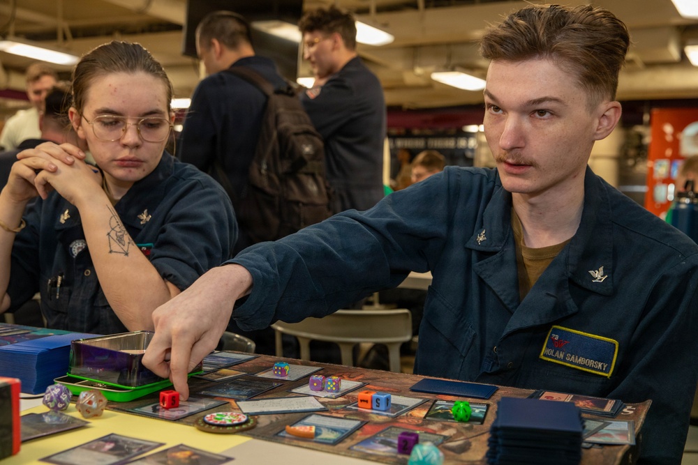 Two U.S. Navy sailors in blue coverall uniforms are playing a trading card game, likely Magic: The Gathering, at a table covered with cards, dice, and game accessories. The male sailor, identified by his name tag as 'Nolan Samborsky,' is reaching across the table to interact with game pieces. The setting appears to be aboard a naval vessel, with other personnel visible in the background.