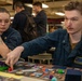 Two U.S. Navy sailors in blue coverall uniforms are playing a trading card game, likely Magic: The Gathering, at a table covered with cards, dice, and game accessories. The male sailor, identified by his name tag as 'Nolan Samborsky,' is reaching across the table to interact with game pieces. The setting appears to be aboard a naval vessel, with other personnel visible in the background.
