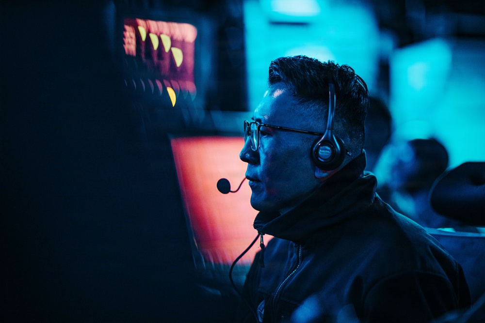 A man wearing glasses and a headset with a boom microphone is seated in front of monitors in a dimly lit environment illuminated by blue and red/orange lighting. He appears focused and engaged, likely communicating with others via the headset. The setting suggests a professional or competitive environment such as an esports event, broadcast booth, or operations center.