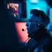 A man wearing glasses and a headset with a boom microphone is seated in front of monitors in a dimly lit environment illuminated by blue and red/orange lighting. He appears focused and engaged, likely communicating with others via the headset. The setting suggests a professional or competitive environment such as an esports event, broadcast booth, or operations center.