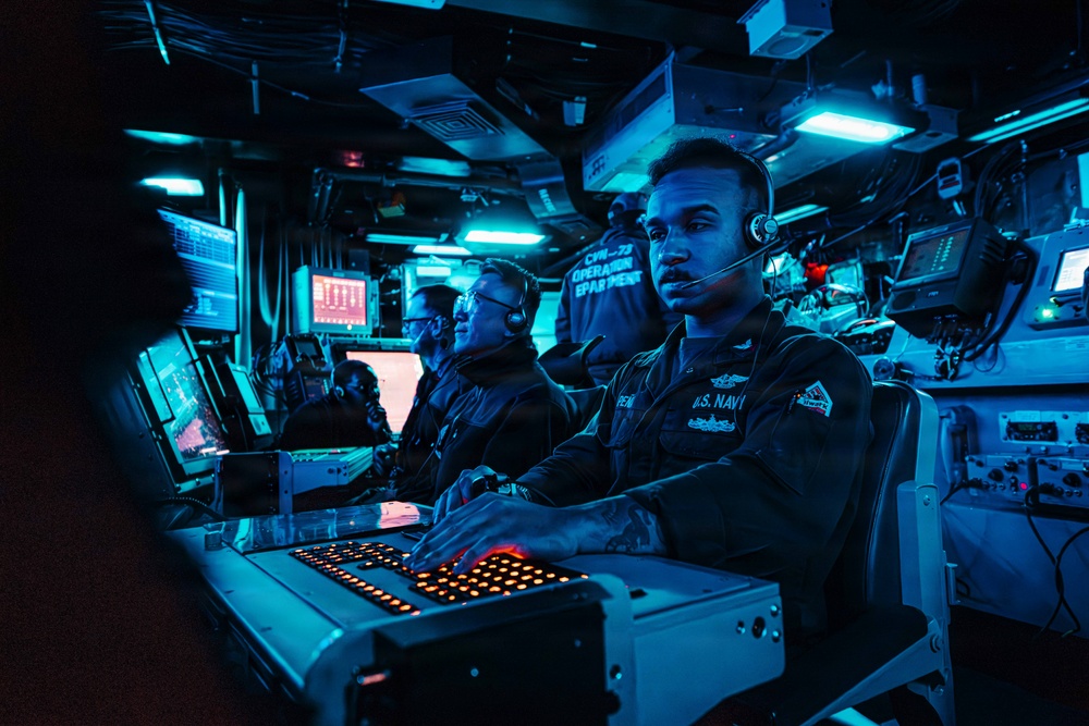 U.S. Navy personnel are stationed at a combat information center (CIC) aboard what appears to be CVN-73 (USS George Washington), operating tactical consoles under blue ambient lighting. Multiple sailors wearing headsets are monitoring screens and operating illuminated control panels in a darkened operations room. The environment is consistent with a ship's operations department during an active watch.