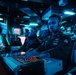 U.S. Navy personnel are stationed at a combat information center (CIC) aboard what appears to be CVN-73 (USS George Washington), operating tactical consoles under blue ambient lighting. Multiple sailors wearing headsets are monitoring screens and operating illuminated control panels in a darkened operations room. The environment is consistent with a ship's operations department during an active watch.