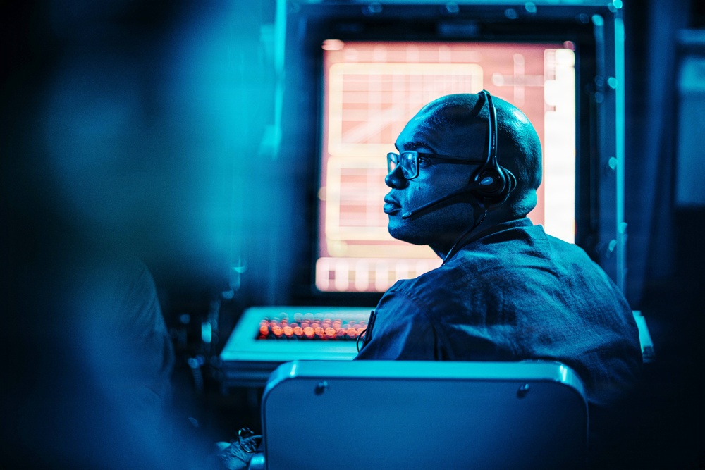 A man wearing glasses and a communication headset is seated at what appears to be a control or monitoring station, illuminated by a strong blue ambient light. He is facing a large glowing monitor displaying what appears to be a grid or data interface with orange/red illumination. The setting suggests a professional operations environment such as a control room, broadcast facility, or command center.