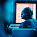 A man wearing glasses and a communication headset is seated at what appears to be a control or monitoring station, illuminated by a strong blue ambient light. He is facing a large glowing monitor displaying what appears to be a grid or data interface with orange/red illumination. The setting suggests a professional operations environment such as a control room, broadcast facility, or command center.