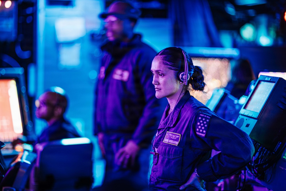 The image depicts what appears to be a military naval operations center or ship bridge, bathed in blue and purple ambient lighting. A female service member in the foreground wearing a headset and dark uniform with visible patches including a US flag patch is the primary focus, appearing alert and attentive. Two additional personnel are visible in the background, also in dark uniforms, surrounded by computer monitors and control equipment.