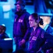 The image depicts what appears to be a military naval operations center or ship bridge, bathed in blue and purple ambient lighting. A female service member in the foreground wearing a headset and dark uniform with visible patches including a US flag patch is the primary focus, appearing alert and attentive. Two additional personnel are visible in the background, also in dark uniforms, surrounded by computer monitors and control equipment.