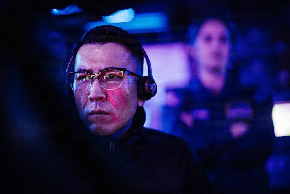 A focused individual wearing glasses and a gaming headset is illuminated by blue and red screen light in a dark environment. The person appears to be intensely concentrating on something off-camera, likely a monitor. A second person wearing a headset is visible but blurred in the background.