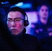 A focused individual wearing glasses and a gaming headset is illuminated by blue and red screen light in a dark environment. The person appears to be intensely concentrating on something off-camera, likely a monitor. A second person wearing a headset is visible but blurred in the background.