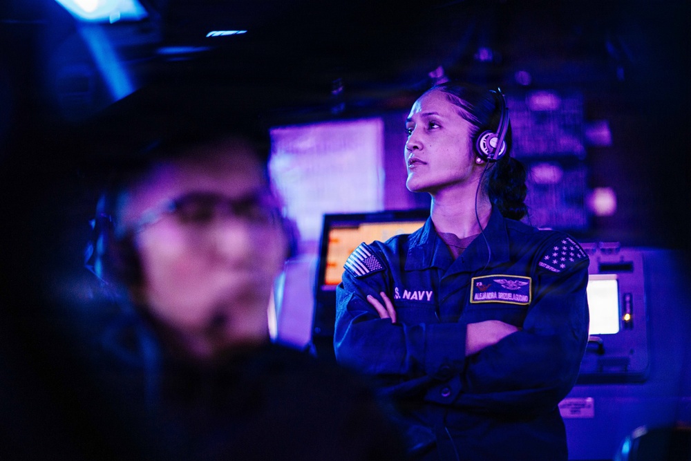 A U.S. Navy sailor wearing a blue uniform and headset stands with arms crossed in what appears to be a ship's combat information center or operations room. The environment is dimly lit with blue/purple lighting, typical of naval operations spaces. A second sailor is visible in the blurred foreground.