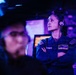 A U.S. Navy sailor wearing a blue uniform and headset stands with arms crossed in what appears to be a ship's combat information center or operations room. The environment is dimly lit with blue/purple lighting, typical of naval operations spaces. A second sailor is visible in the blurred foreground.