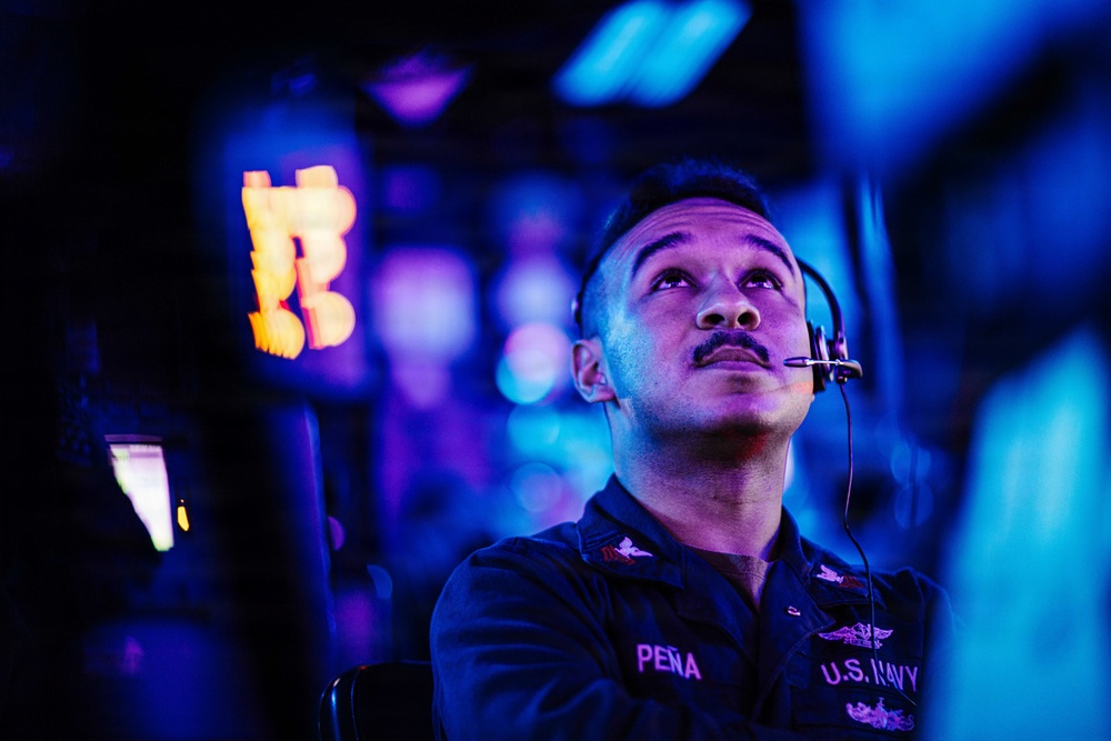 A U.S. Navy sailor named Peña is seated at what appears to be a combat information center or operations room aboard a naval vessel. He is wearing a headset and looking upward, likely monitoring displays or receiving communications. The environment is illuminated with blue and purple lighting typical of shipboard operations centers.
