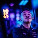 A U.S. Navy sailor named Peña is seated at what appears to be a combat information center or operations room aboard a naval vessel. He is wearing a headset and looking upward, likely monitoring displays or receiving communications. The environment is illuminated with blue and purple lighting typical of shipboard operations centers.