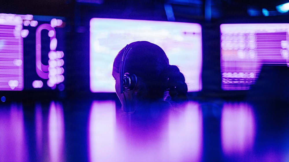 A person wearing headphones is seated in front of multiple illuminated monitors in a darkened room bathed in purple and blue lighting. The scene suggests a professional monitoring, control room, or gaming environment. The silhouetted figure faces away from the camera, focused on the screens ahead.