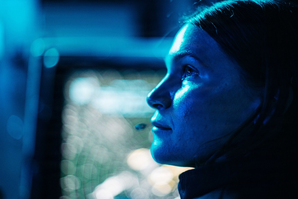 A close-up profile shot of a person illuminated by intense blue light, appearing to gaze upward or into the distance. The image has a cinematic, dramatic quality with a shallow depth of field and bokeh background lights. The subject's face shows visible moisture or sweat, and there appears to be a tear on the cheek.