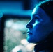 A close-up profile shot of a person illuminated by intense blue light, appearing to gaze upward or into the distance. The image has a cinematic, dramatic quality with a shallow depth of field and bokeh background lights. The subject's face shows visible moisture or sweat, and there appears to be a tear on the cheek.
