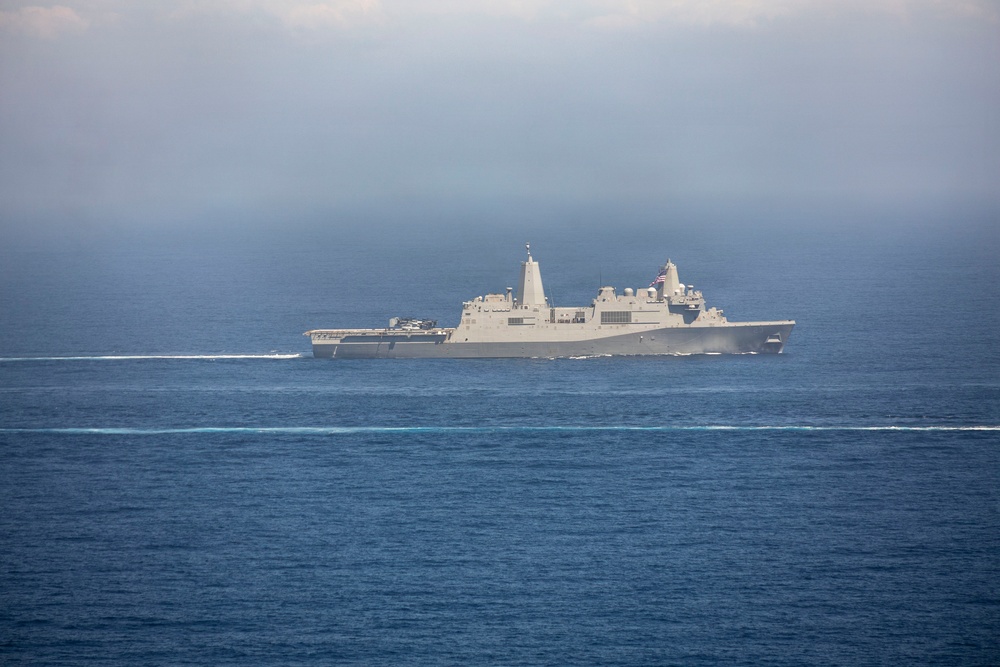 A large gray military naval vessel, consistent with a US Navy amphibious transport dock (LPD) ship, is underway on open blue ocean waters. The ship is traveling at moderate speed, visible by the wake it generates. An American flag is visible on the vessel's mast area, and atmospheric haze partially obscures the background sky.