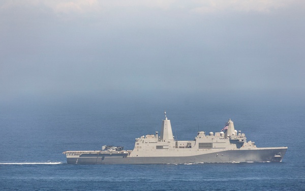 A large gray military naval vessel, consistent with a US Navy amphibious transport dock (LPD) ship, is underway on open blue ocean waters. The ship is traveling at moderate speed, visible by the wake it generates. An American flag is visible on the vessel's mast area, and atmospheric haze partially obscures the background sky.