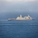 A large gray military naval vessel, consistent with a US Navy amphibious transport dock (LPD) ship, is underway on open blue ocean waters. The ship is traveling at moderate speed, visible by the wake it generates. An American flag is visible on the vessel's mast area, and atmospheric haze partially obscures the background sky.
