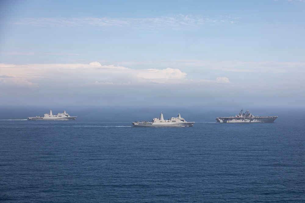 Three large gray naval vessels are sailing in formation across open blue ocean waters. The ships appear to be US Navy amphibious warfare vessels, including what appears to be an amphibious assault ship on the right and two amphibious transport dock ships. The image is taken from an elevated aerial perspective under a partly cloudy sky.