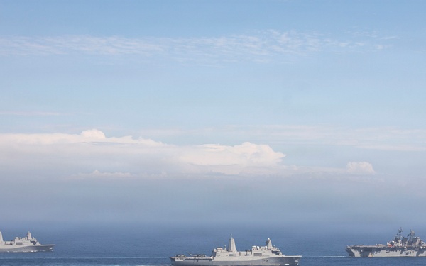 Three large gray naval vessels are sailing in formation across open blue ocean waters. The ships appear to be US Navy amphibious warfare vessels, including what appears to be an amphibious assault ship on the right and two amphibious transport dock ships. The image is taken from an elevated aerial perspective under a partly cloudy sky.