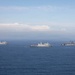 Three large gray naval vessels are sailing in formation across open blue ocean waters. The ships appear to be US Navy amphibious warfare vessels, including what appears to be an amphibious assault ship on the right and two amphibious transport dock ships. The image is taken from an elevated aerial perspective under a partly cloudy sky.