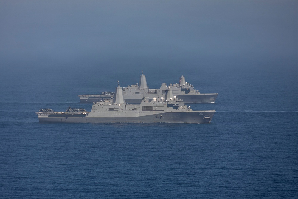 The image shows multiple U.S. Navy warships sailing in formation on open ocean. At least three vessels are visible, including what appears to be San Antonio-class amphibious transport dock ships and a larger vessel in the background that resembles an aircraft carrier or amphibious assault ship. The ships are painted in standard U.S. Navy haze gray and are underway in calm seas.