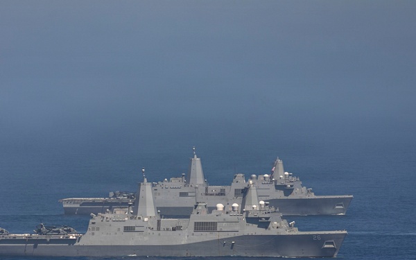 The image shows multiple U.S. Navy warships sailing in formation on open ocean. At least three vessels are visible, including what appears to be San Antonio-class amphibious transport dock ships and a larger vessel in the background that resembles an aircraft carrier or amphibious assault ship. The ships are painted in standard U.S. Navy haze gray and are underway in calm seas.