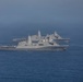 The image shows multiple U.S. Navy warships sailing in formation on open ocean. At least three vessels are visible, including what appears to be San Antonio-class amphibious transport dock ships and a larger vessel in the background that resembles an aircraft carrier or amphibious assault ship. The ships are painted in standard U.S. Navy haze gray and are underway in calm seas.