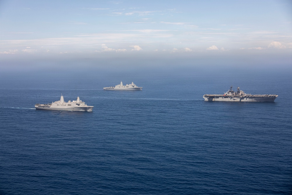 An aerial photograph shows three U.S. Navy warships sailing in formation across open blue ocean waters. The formation includes what appears to be an amphibious assault ship (largest vessel, right), and two San Antonio-class amphibious transport dock ships (LPD). The ships are underway in calm seas under a partly cloudy sky, suggesting a naval exercise or transit operation.