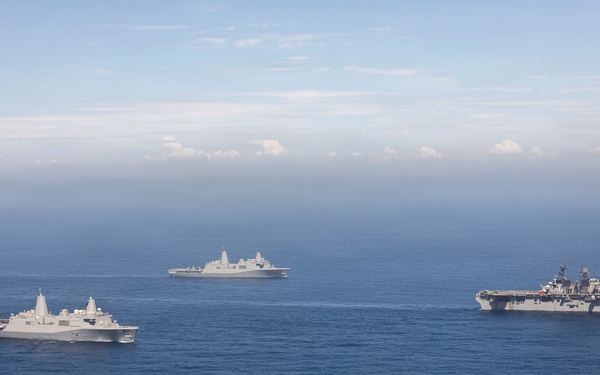 An aerial photograph shows three U.S. Navy warships sailing in formation across open blue ocean waters. The formation includes what appears to be an amphibious assault ship (largest vessel, right), and two San Antonio-class amphibious transport dock ships (LPD). The ships are underway in calm seas under a partly cloudy sky, suggesting a naval exercise or transit operation.