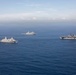An aerial photograph shows three U.S. Navy warships sailing in formation across open blue ocean waters. The formation includes what appears to be an amphibious assault ship (largest vessel, right), and two San Antonio-class amphibious transport dock ships (LPD). The ships are underway in calm seas under a partly cloudy sky, suggesting a naval exercise or transit operation.