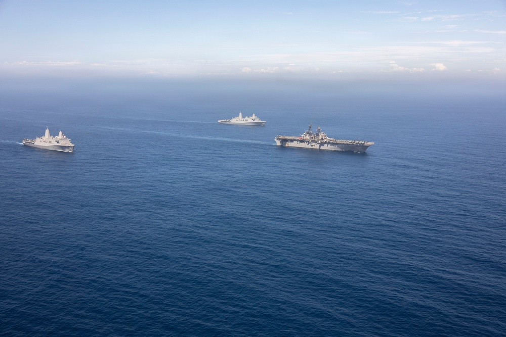 An aerial photograph shows three naval vessels sailing in formation across open blue ocean waters. The formation includes what appears to be a large amphibious assault ship or aircraft carrier on the right, a smaller amphibious transport dock ship on the left, and a third vessel in the background center. The ships appear to be conducting a joint naval exercise or transit operation.
