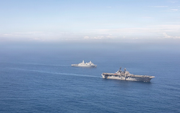 An aerial photograph shows three naval vessels sailing in formation across open blue ocean waters. The formation includes what appears to be a large amphibious assault ship or aircraft carrier on the right, a smaller amphibious transport dock ship on the left, and a third vessel in the background center. The ships appear to be conducting a joint naval exercise or transit operation.