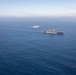 An aerial photograph shows three naval vessels sailing in formation across open blue ocean waters. The formation includes what appears to be a large amphibious assault ship or aircraft carrier on the right, a smaller amphibious transport dock ship on the left, and a third vessel in the background center. The ships appear to be conducting a joint naval exercise or transit operation.
