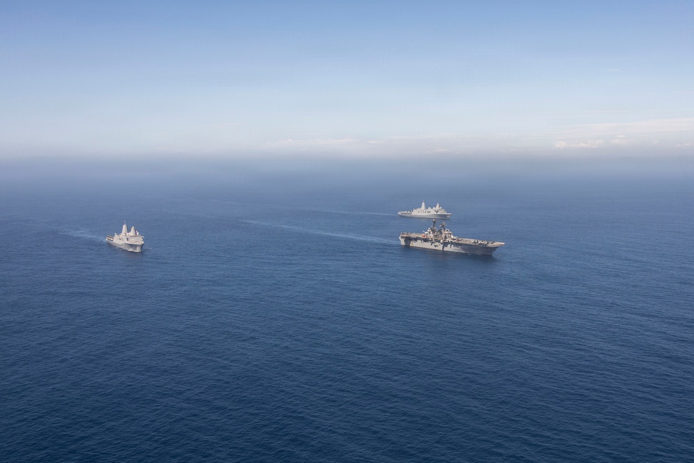 An aerial photograph shows three naval vessels sailing in formation across open blue ocean waters. The formation includes one large amphibious assault ship or aircraft carrier in the foreground, one smaller vessel to the left, and a third ship visible in the background. The ships appear to be underway in calm sea conditions under a hazy blue sky.