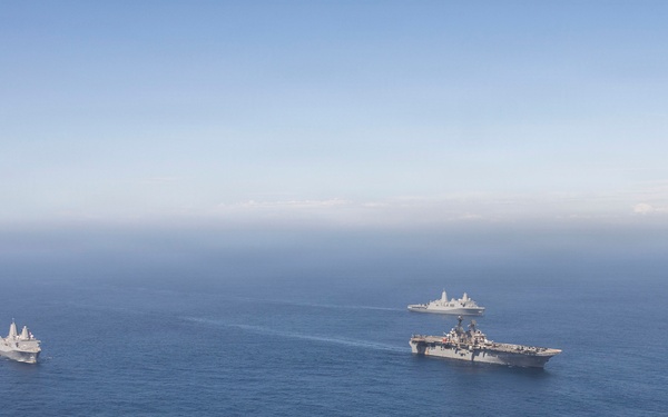 An aerial photograph shows three naval vessels sailing in formation across open blue ocean waters. The formation includes one large amphibious assault ship or aircraft carrier in the foreground, one smaller vessel to the left, and a third ship visible in the background. The ships appear to be underway in calm sea conditions under a hazy blue sky.
