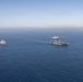 An aerial photograph shows three naval vessels sailing in formation across open blue ocean waters. The formation includes one large amphibious assault ship or aircraft carrier in the foreground, one smaller vessel to the left, and a third ship visible in the background. The ships appear to be underway in calm sea conditions under a hazy blue sky.