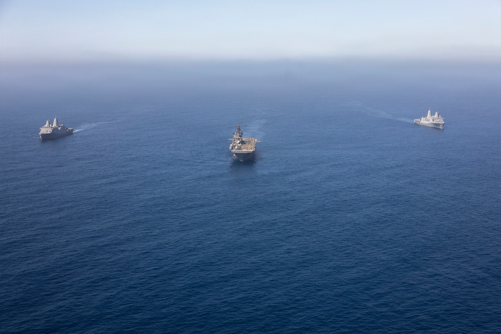An aerial photograph shows three naval vessels sailing in formation across open blue ocean water. The center ship appears to be a large amphibious assault ship or helicopter carrier, flanked by two smaller warships on either side. All three vessels are underway, producing visible wakes as they travel in the same direction.