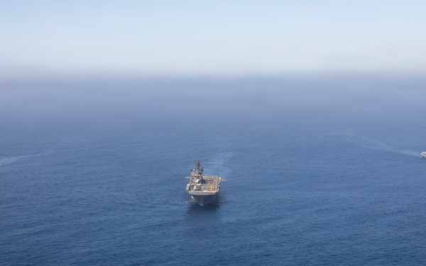 An aerial photograph shows three naval vessels sailing in formation across open blue ocean water. The center ship appears to be a large amphibious assault ship or helicopter carrier, flanked by two smaller warships on either side. All three vessels are underway, producing visible wakes as they travel in the same direction.