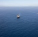 An aerial photograph shows three naval vessels sailing in formation across open blue ocean water. The center ship appears to be a large amphibious assault ship or helicopter carrier, flanked by two smaller warships on either side. All three vessels are underway, producing visible wakes as they travel in the same direction.